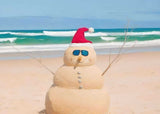 A sand snowman with a Santa hat and sunglasses stands on a sunny beach with turquoise waves in the background.